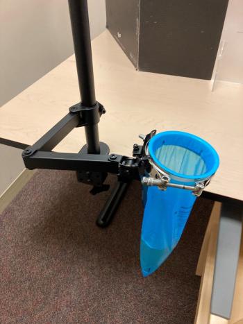 Photos depict an adjustable metal device that allows for hands-free collection of taste testing expectorants in emesis bags. The device attaches to the tabletop with a clamp and has one arm holding the emesis bag open. The height of the bag from the floor is adjustable.
